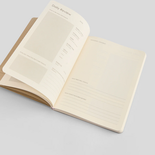 An open journal with off-white, sectioned pages titled 'Daily Review'. Sections include space to write goals, practice self-care, and reflect on the day.