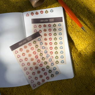 Two sheets of decorative fruit stickers with a dark header are laid on an open notebook, alongside a roll of tape and an orange-handled tool, all placed on a textured mustard-yellow surface with dappled sunlight.